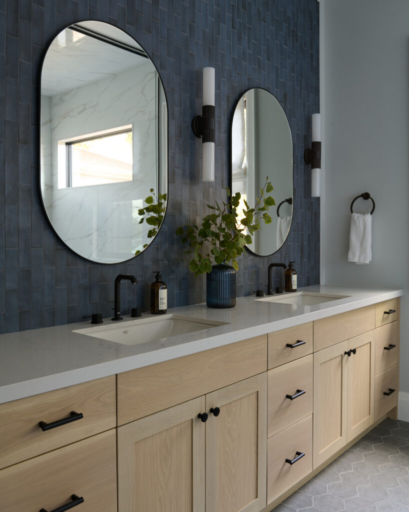 Primary bathroom with double vanity in light wood, navy stone backsplash to ceiling, black hardware, black-trim mirrors and dark grey hexagon tile floor, designed by a Toronto-area interior design studio Erin Interiors.