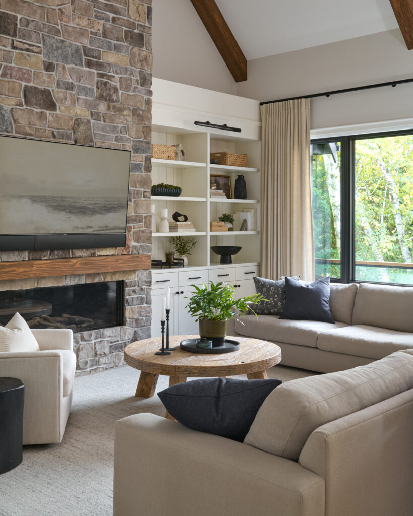 Custom living room with floor-to-ceiling stone fireplace, built-in shelving with open display and closed storage, black-trimmed window with linen drapery, round coffee table and neutral rug, designed by a Toronto, Ontario interior designer.