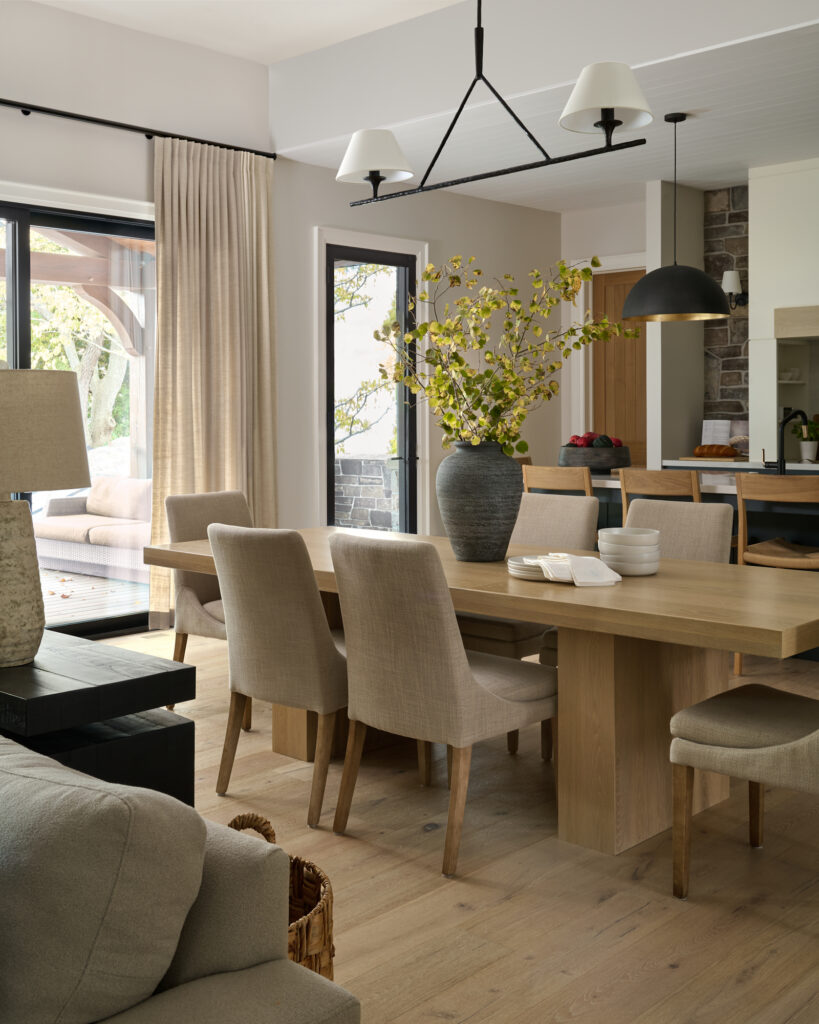 Open-concept dining area with light wood table in front of black-trim patio doors, transitioning between living room and kitchen with island, bar stools and black pendant lighting, designed by a Toronto interior design studio.