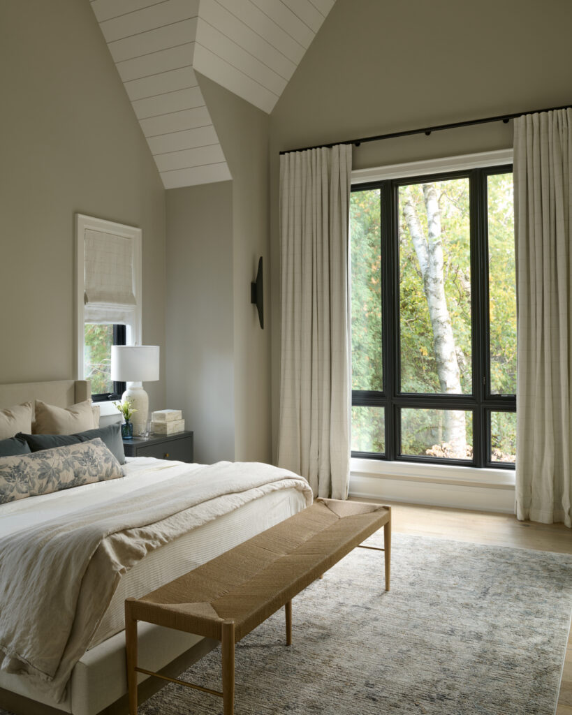 Bright primary bedroom with vaulted ceilings, king bed and woven bench, warm grey walls, light wood flooring, neutral linens and custom curtains on black-trim window, designed by a Toronto-area interior design studio Erin Interiors.