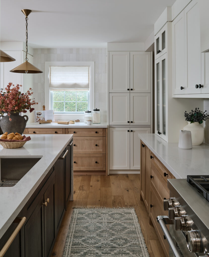 This farmhouse kitchen design blends light oak cabinets and flooring with a contrasting dark island for a warm, layered kitchen aesthetic. Brass pendant lights hang above the island, while pale tile in soft white and greige tones offers timeless kitchen backsplash ideas. Glass upper cabinets, a stainless steel stove, appliance barn, and neutral roman shade add charm and function. Save this inviting kitchen interior for classic kitchen design ideas that feel bright and grounded.