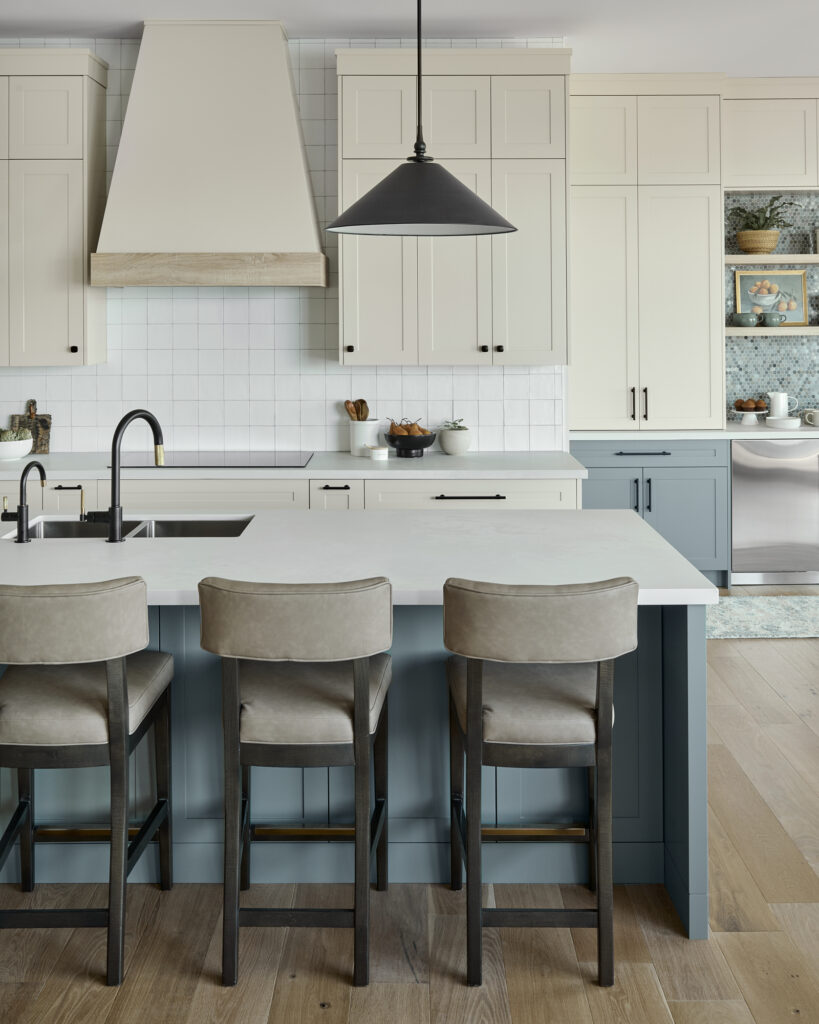 A modern kitchen renovation by Erin Interiors, an interior design team in Whiby, Ontario, features lower cabinetry in Duxbury Gray by Benjamin Moore and upper cabinetry in Edgecomb Grey by Benjamin Moore. There are counter height chairs at the island with a black pendant light over the kitchen island.