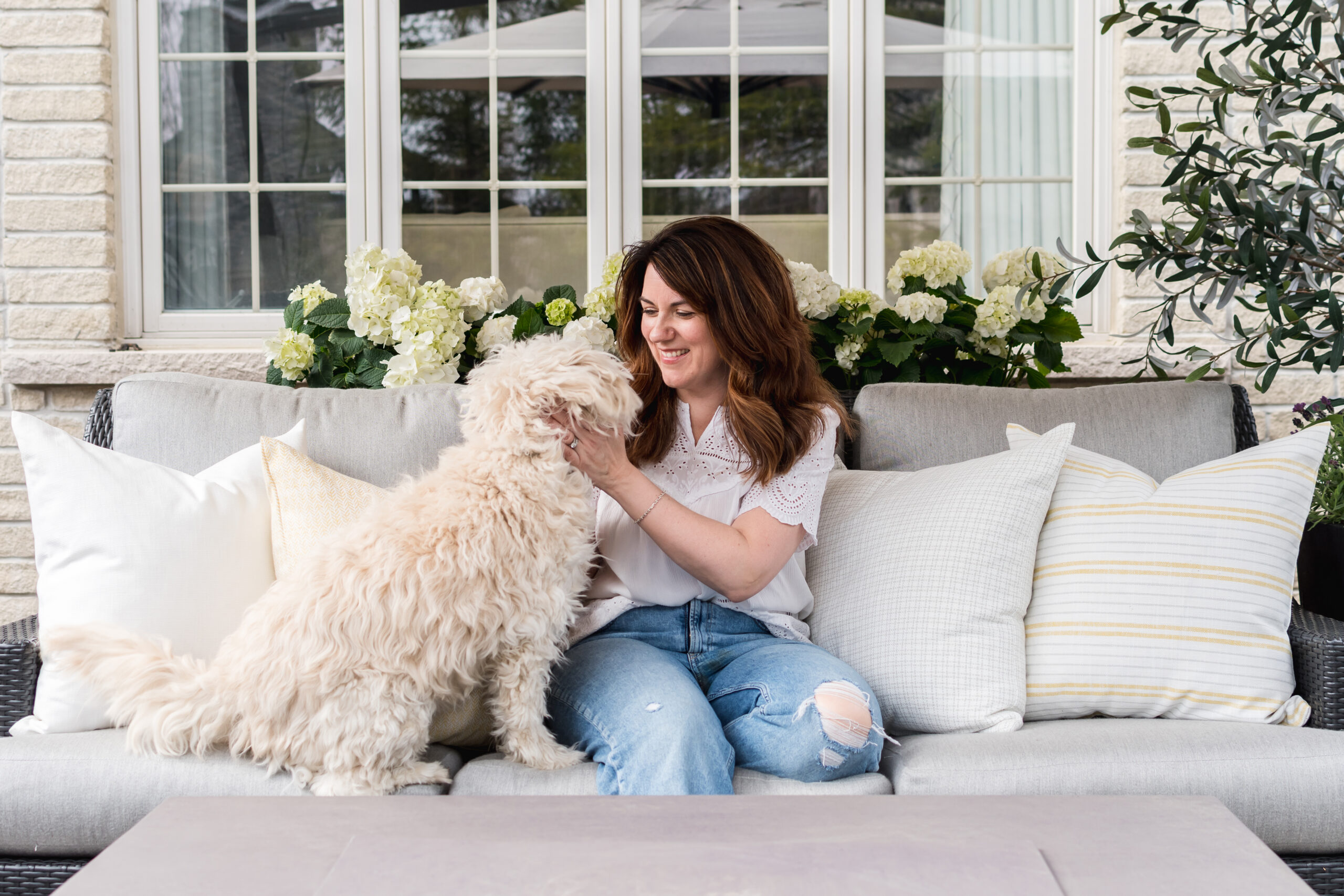 Whitby interior designer Erin Jones of Erin Interiors sits on a grey outdoor sofa with her white shaggy dog. There are yellow and cream coloured outdoor pillows behind them.