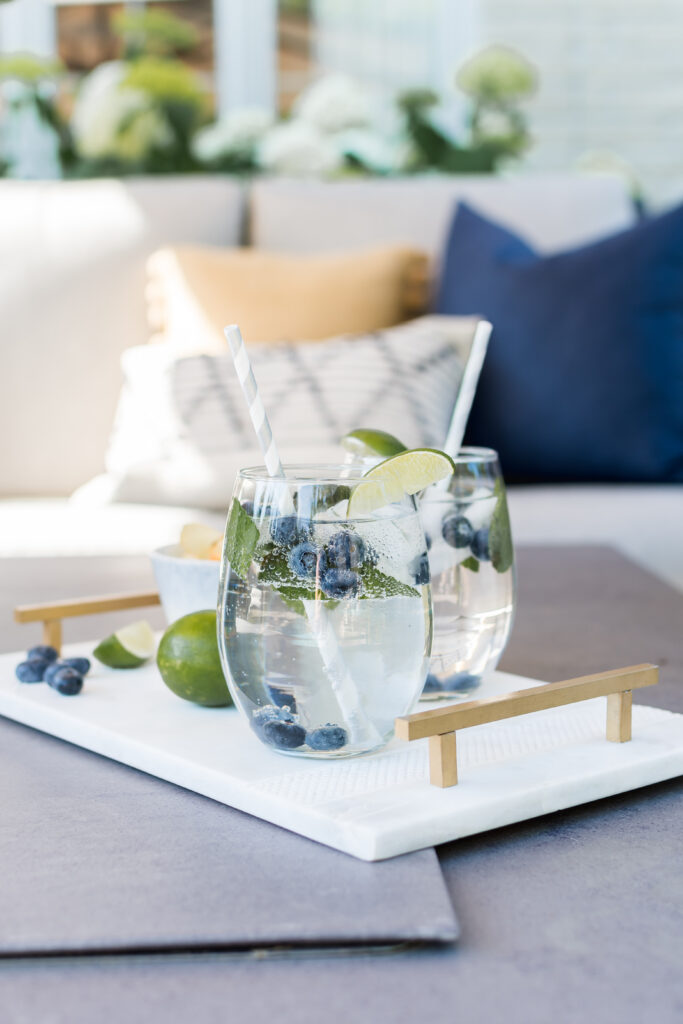 Two stemless wine glasses with blueberry mojitos are on a white tray with gold handles. The background includes an outdoor sofa with navy blue, yellow and white outdoor pillows.