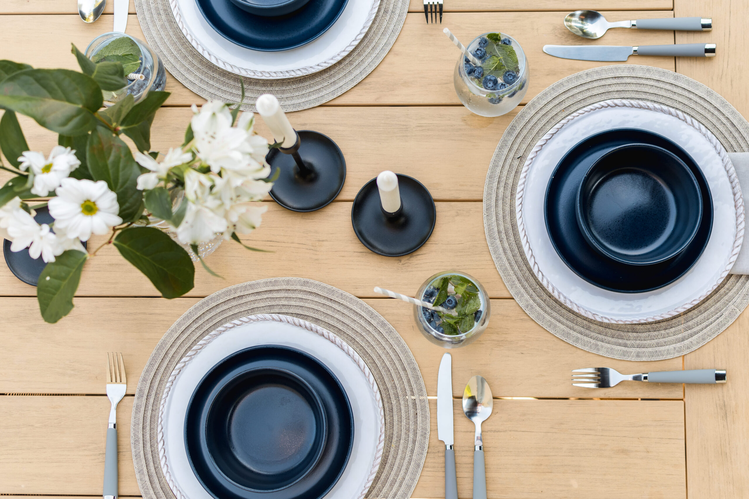 An outdoor dining table is shown from above with woven round placemats and navy blue dishes set for a meal. The table is a light wood tone and there are candlesticks and a vase of white flowers as well as blueberry mojitos ready to be enjoyed.