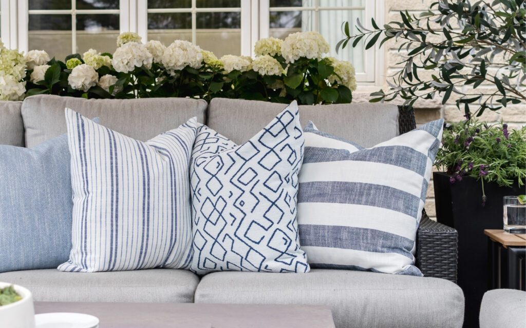 A charcoal grey outdoor conversation set with grey back and seat cushions is adorned with patterned outdoor throw pillows in various blue and white patterns. There are potted white hydrangeas visible behind the outdoor sofa and the table in front of it has a decorative bowl of moss and a vase of delicate purple flowers.