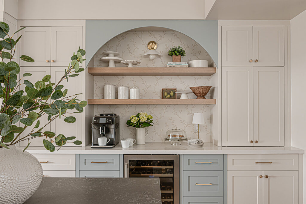 Cream kitchen with an arched coffee bar nook in sage blue, floating wood shelves, brass fixtures, and a built-in wine fridge.