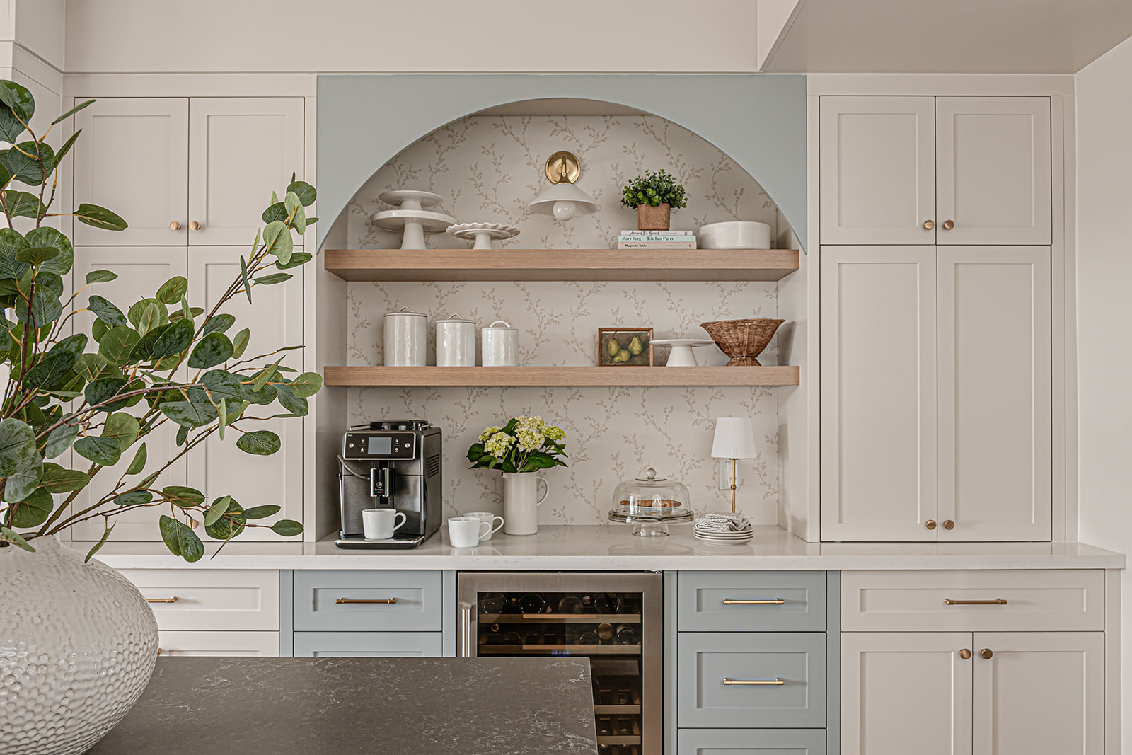 Cream kitchen with an arched coffee bar nook in sage blue, floating wood shelves, brass fixtures, and a built-in wine fridge.
