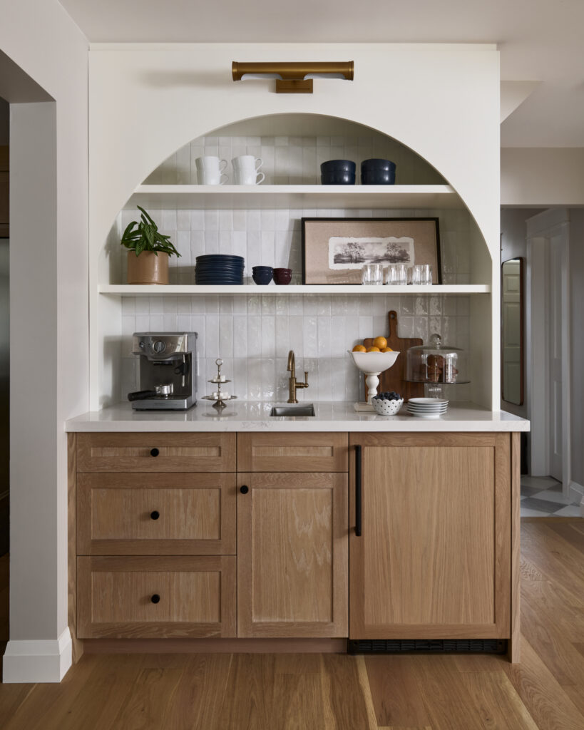 A thoughtfully designed coffee bar doubles as a wet bar in the kitchen, complete with a mini sink, creamy white shelving, and light oak cabinetry with black hardware. The arched wall detail & vertical tile add architectural charm. If you're looking for refined coffee bar ideas and timeless wet bar ideas with sink, look no further than this Toronto interior design project by Erin Interiors, completed as part of a kitchen renovation. It's the perfect coffee bar inspiration for stylish mornings.