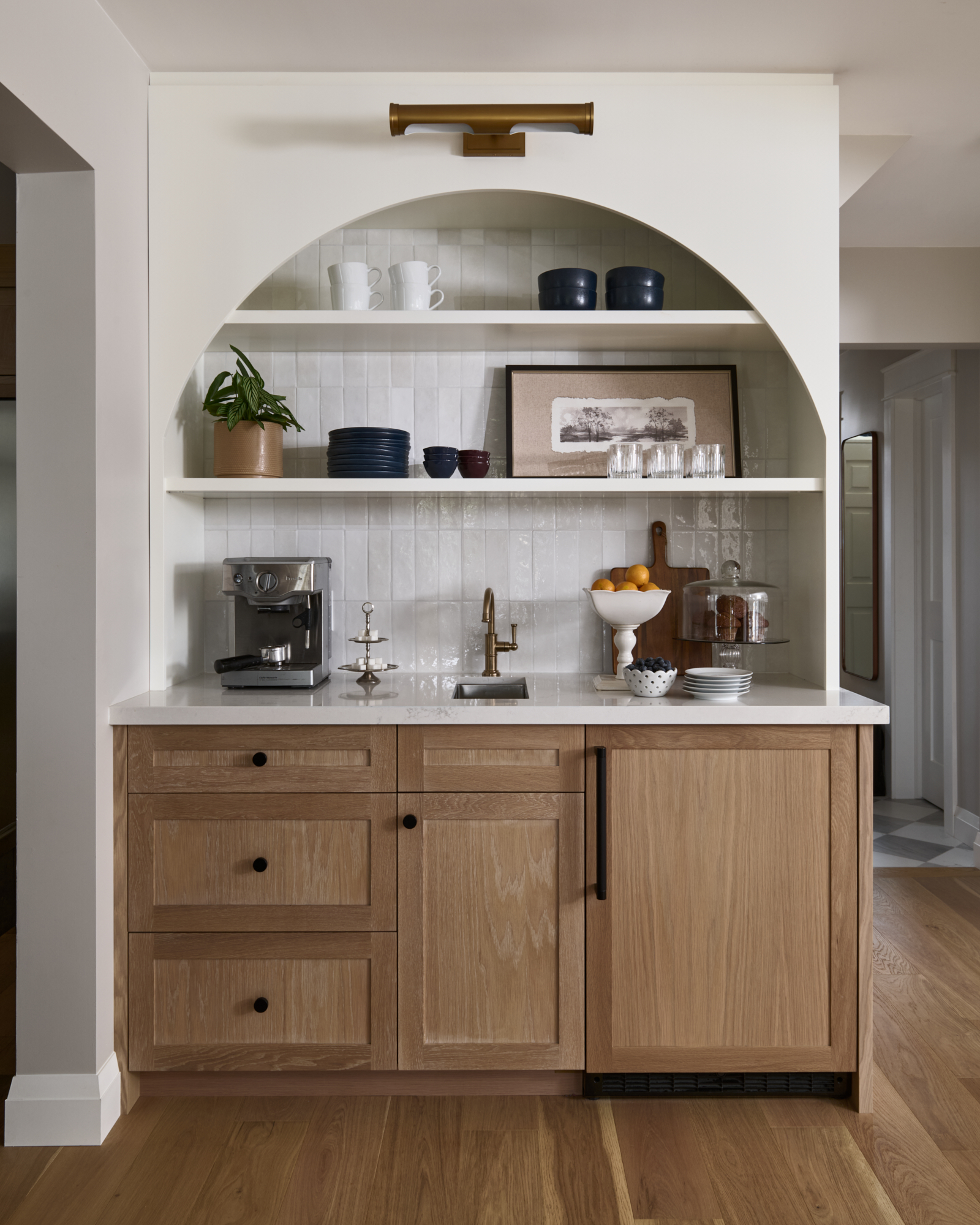 A thoughtfully designed coffee bar doubles as a wet bar in the kitchen, complete with a mini sink, creamy white shelving, and light oak cabinetry with black hardware. The arched wall detail & vertical tile add architectural charm. If you're looking for refined coffee bar ideas and timeless wet bar ideas with sink, look no further than this Toronto interior design project by Erin Interiors, completed as part of a kitchen renovation. It's the perfect coffee bar inspiration for stylish mornings.