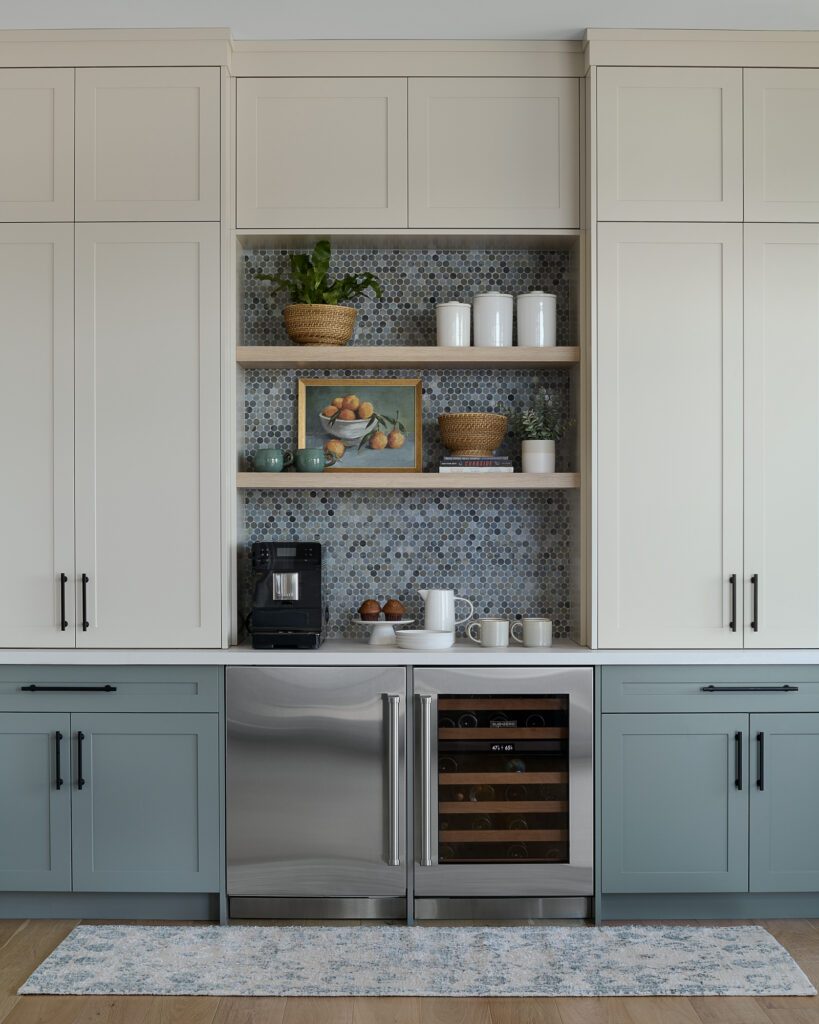 This kitchen coffee bar and beverage station has a double door bar fridge under the counter with a coffee maker and mugs sitting on the white countertop. Above the coffee maker there are two open shelves with dishes and decor on them. The backsplash is penny tile in blue tones.