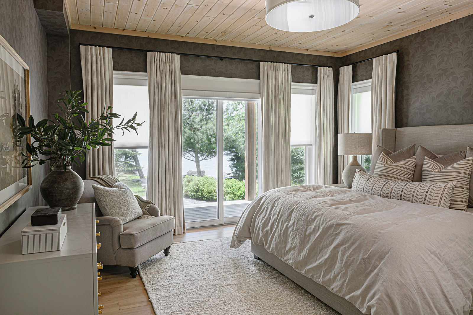 Primary bedroom suite in an Ontario cottage with a pine plank ceiling, moody botanical wallpaper, an upholstered bed, and windows overlooking the lake with linen drapery.