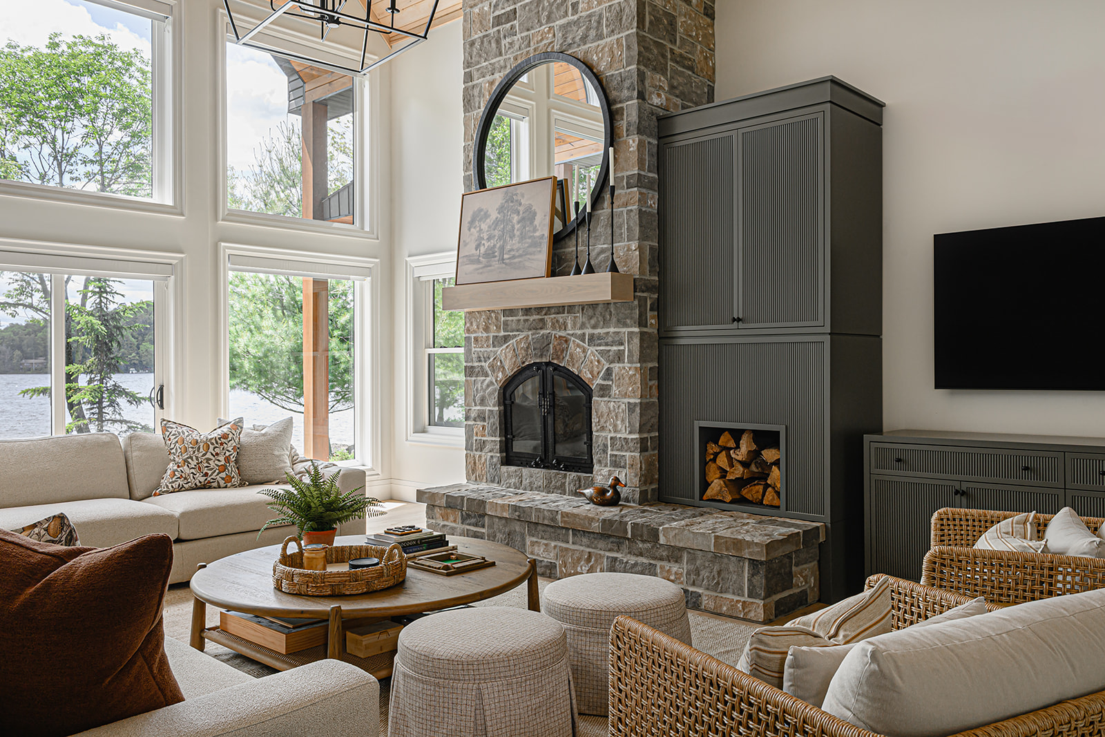 Ontario cottage family room with a floor-to-ceiling stone fireplace, two-storey lake-view windows, and cream sofas paired with rattan armchairs.