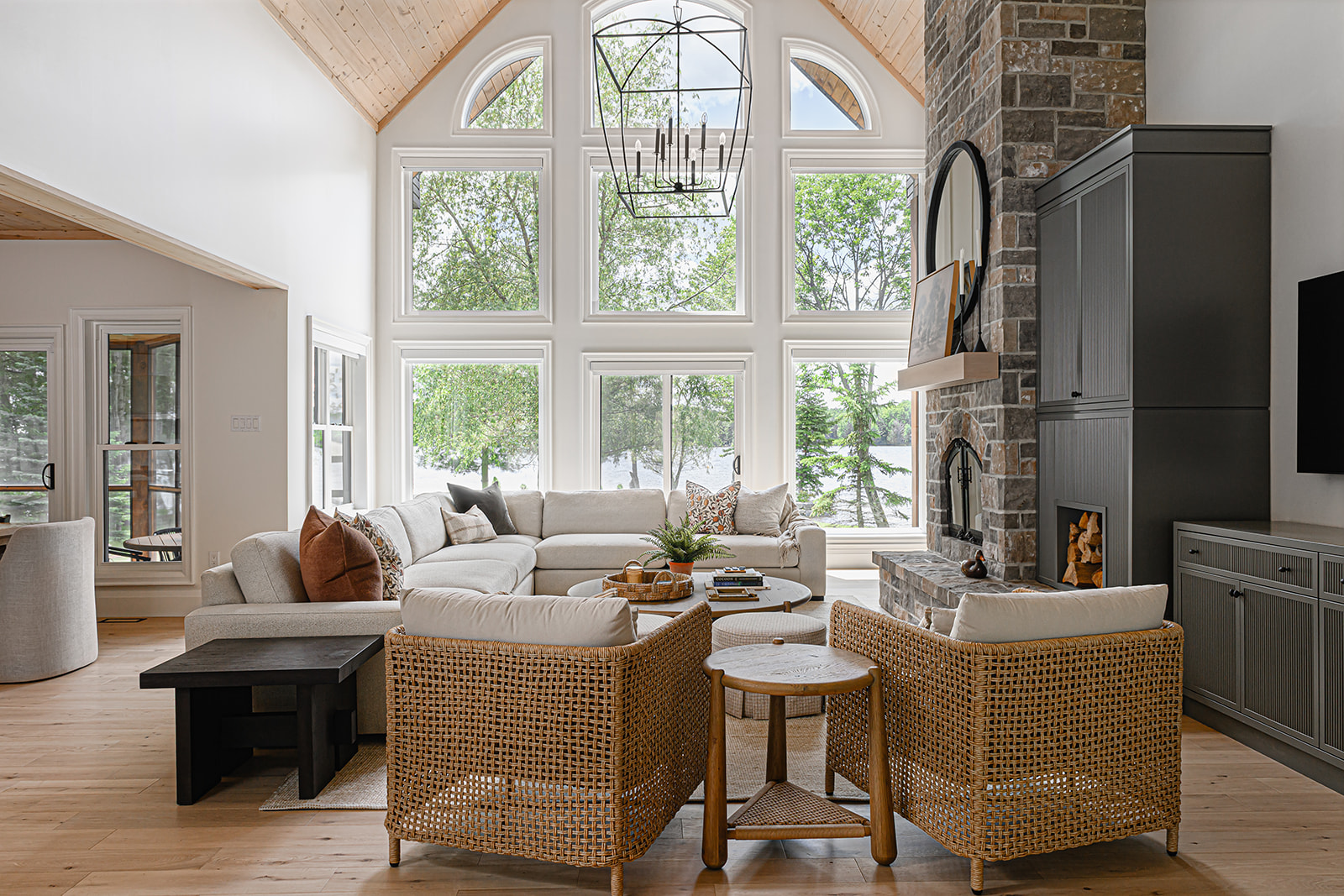 Family room in an Ontario cottage with a double-height window wall, a black lantern chandelier, a cream sectional, and woven armchairs overlooking the lake.