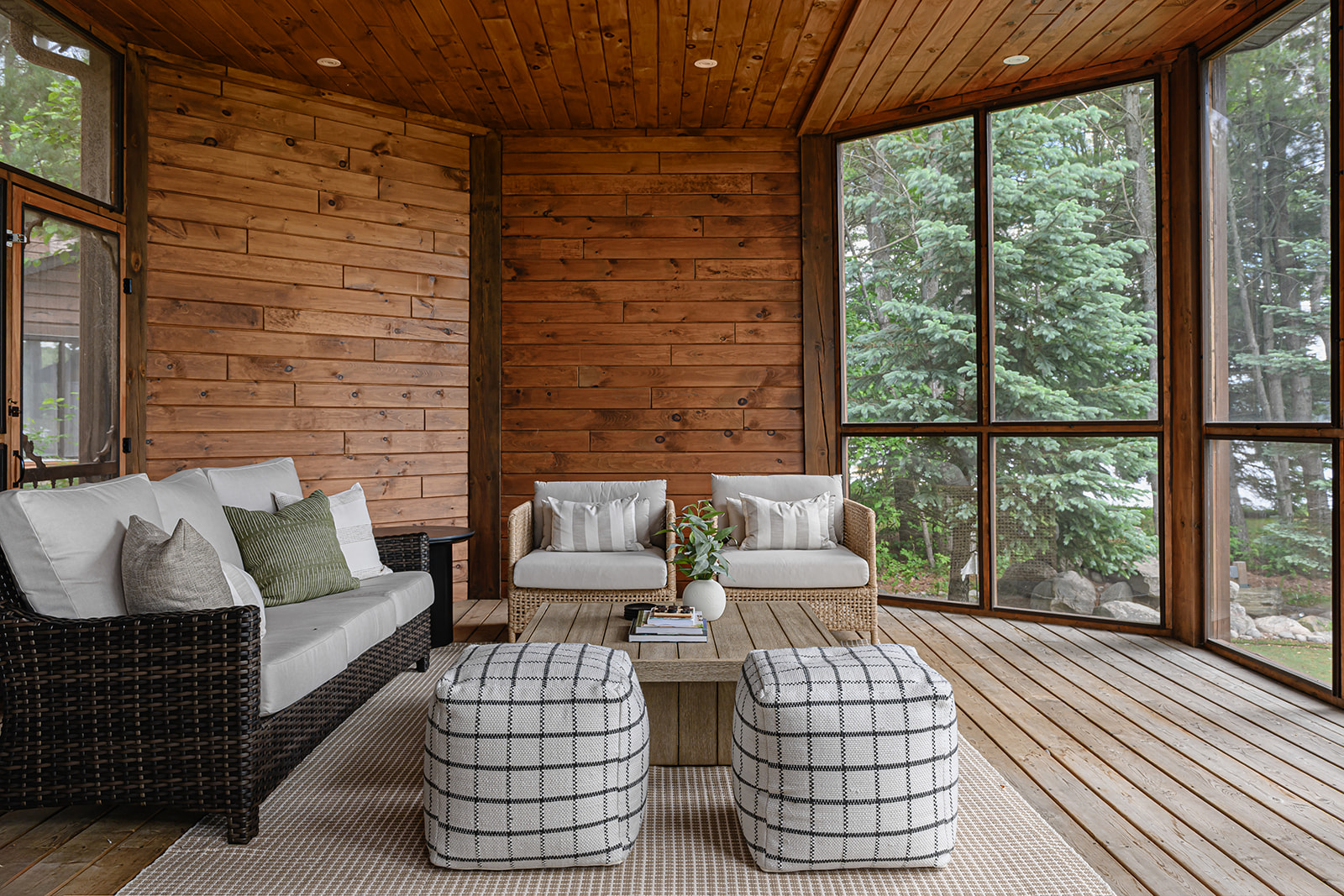Screened porch in an Ontario cottage with wood plank walls and ceiling, a dark wicker sofa, rattan chairs, and checked poufs facing the trees.