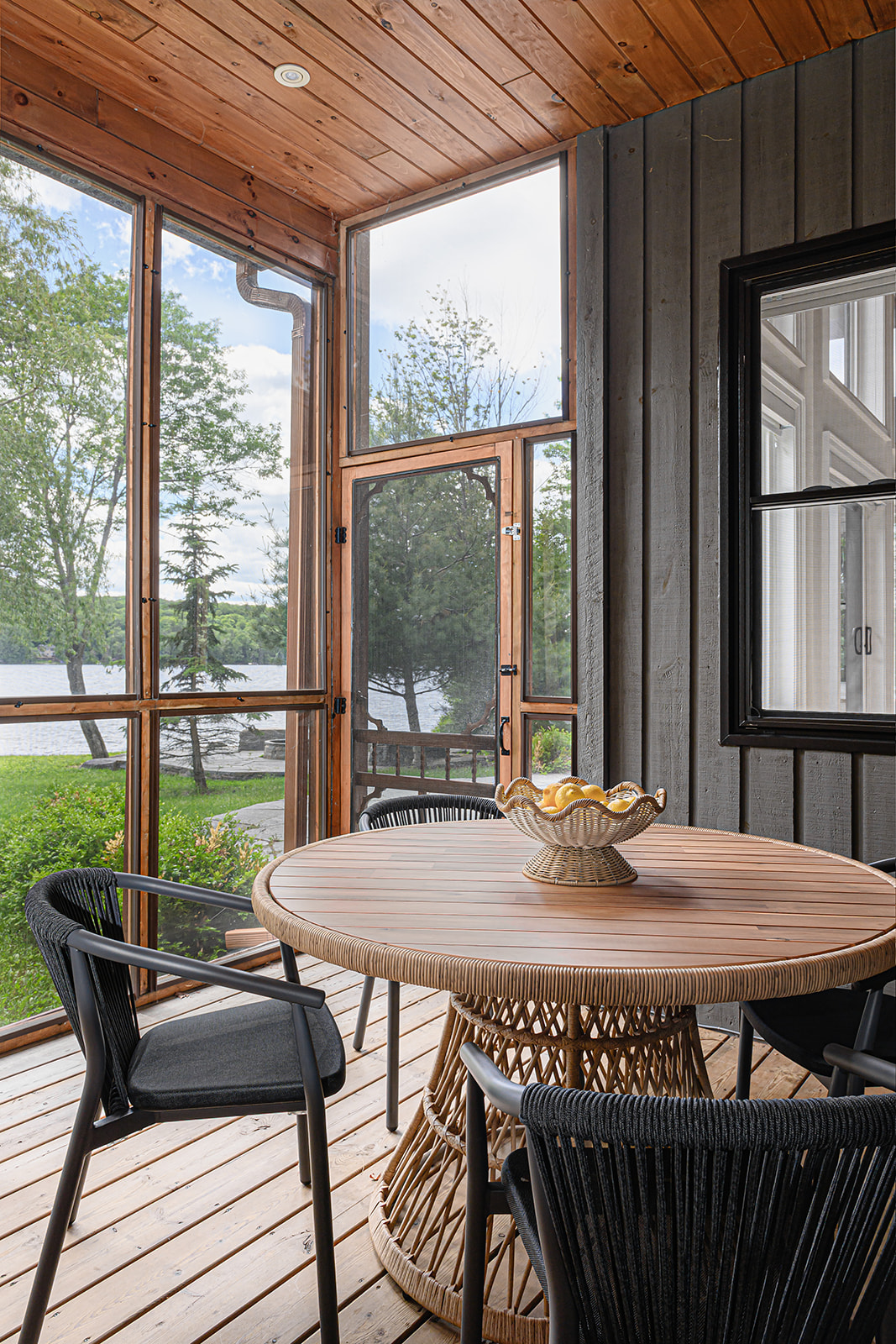 Dining area on an Ontario cottage screened porch with a round rattan table, black woven chairs, a wood plank ceiling, and a lake view through the screens.