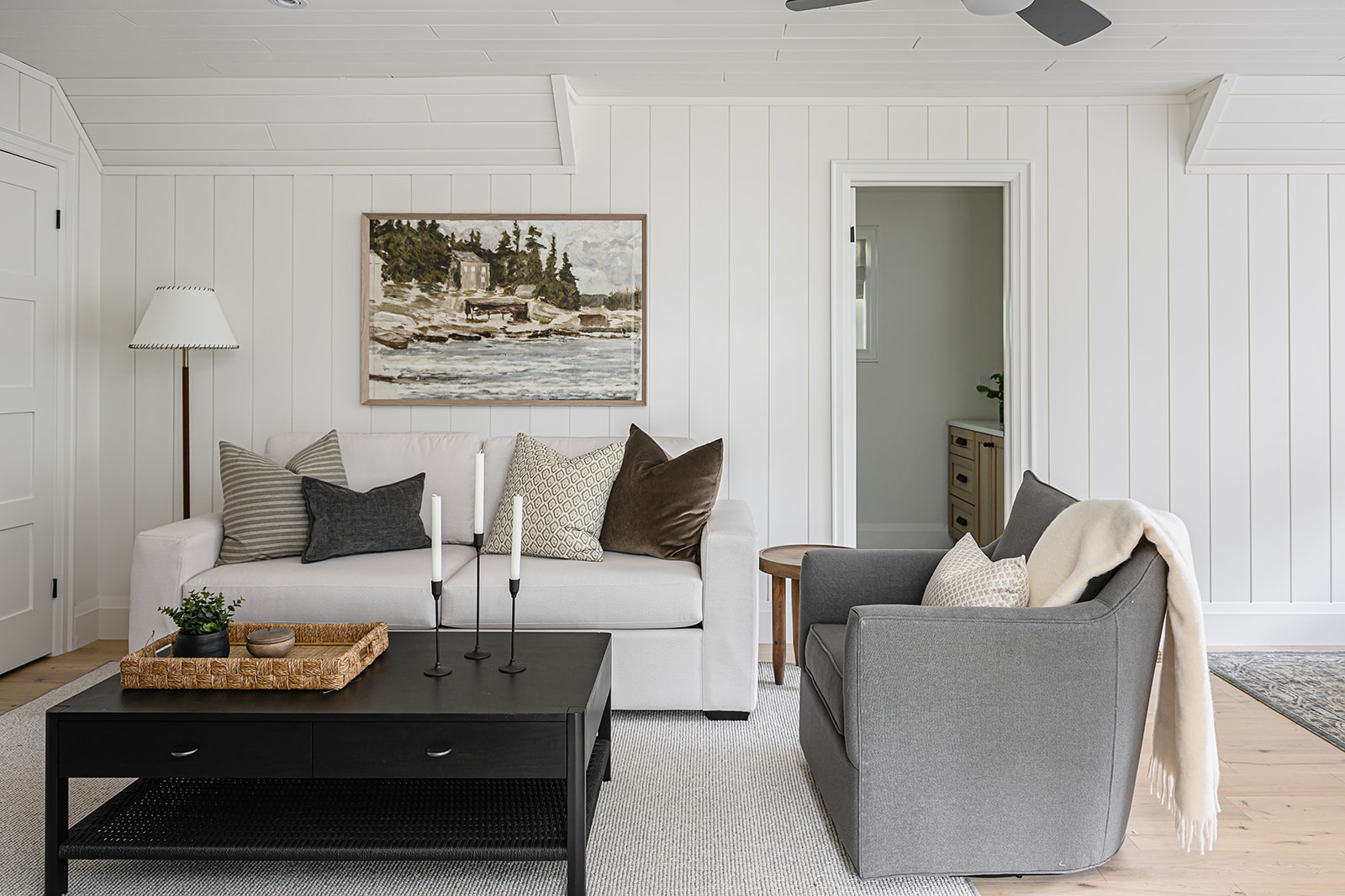 Sitting area in an Ontario cottage bunkie with white shiplap walls, a cream sofa below a framed landscape painting, a grey armchair, and a black coffee table.
